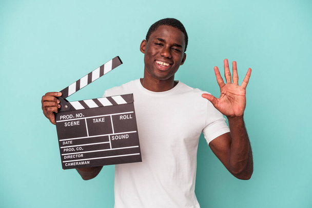 młody Afroamerykanin trzyma klapperboard odizolowany na niebieskim tle uśmiechnięty radosny pokazując numer pięć z palcami. - Zdjęcie, obraz