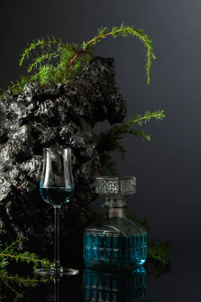 Blue gin on a black reflective background. Juniper branches and grey stone are in the background. - Φωτογραφία, εικόνα