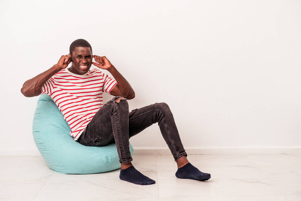 Νεαρός Αφροαμερικάνος κάθεται σε μια ρουφηξιά απομονωμένος σε λευκό φόντο καλύπτοντας τα αυτιά με τα χέρια. - Φωτογραφία, εικόνα