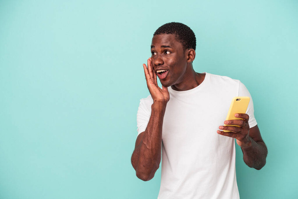 Młody Afroamerykanin trzymający telefon komórkowy na niebieskim tle mówi sekretną wiadomość o gorącym hamowaniu i odwraca wzrok. - Zdjęcie, obraz