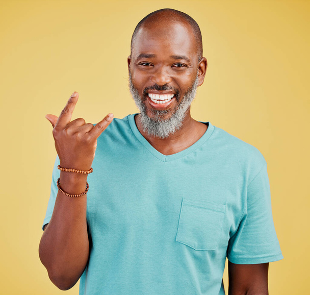 Happy mature african man expressing a rock and roll symbol gesture with his hands against a yellow studio background. Carefree, cool and confident black man making horn gesture to show good vibes. - Foto, Bild