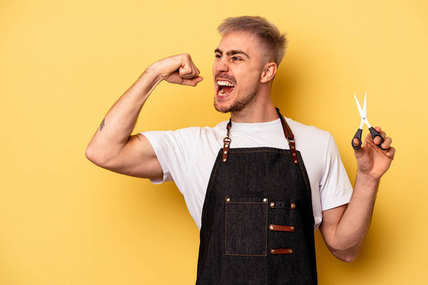 Young caucasian hairdresser man holding scissors isolated on yellow background raising fist after a victory, winner concept. - Fotografie, Obrázek