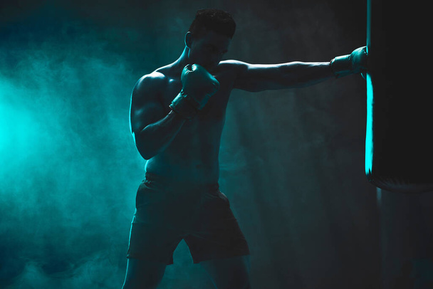 Silhouette of an unrecognizable boxer man sparring with a punching bag in a dark gym. Male athlete working out in preparation for his upcoming fight. Young martial artist training and practicing. - 写真・画像