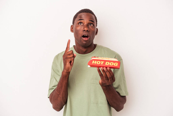 Junger afroamerikanischer Mann isst einen Hot Dog isoliert auf weißem Hintergrund und zeigt mit geöffnetem Mund auf den Kopf. - Foto, Bild