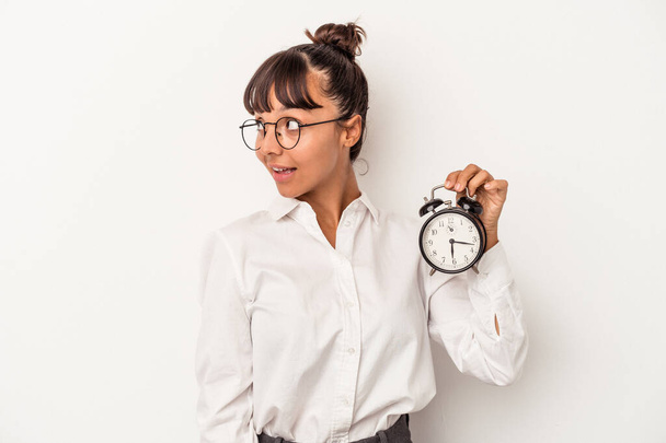 Joven mujer de negocios de raza mixta sosteniendo un reloj despertador aislado sobre fondo blanco se ve a un lado sonriente, alegre y agradable. - Foto, imagen