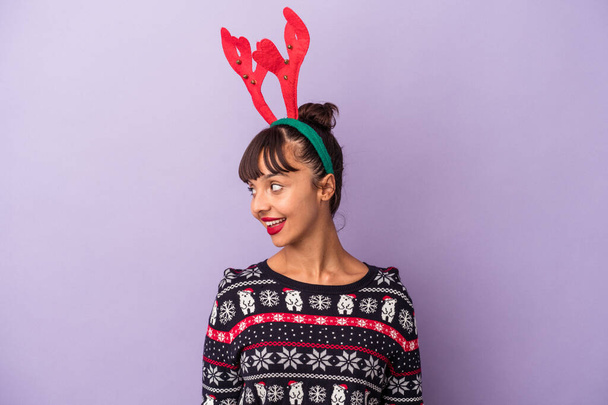 Jonge gemengde ras vrouw met rendieren hoed vieren Kerstmis geïsoleerd op paarse achtergrond kijkt opzij glimlachend, vrolijk en aangenaam. - Foto, afbeelding