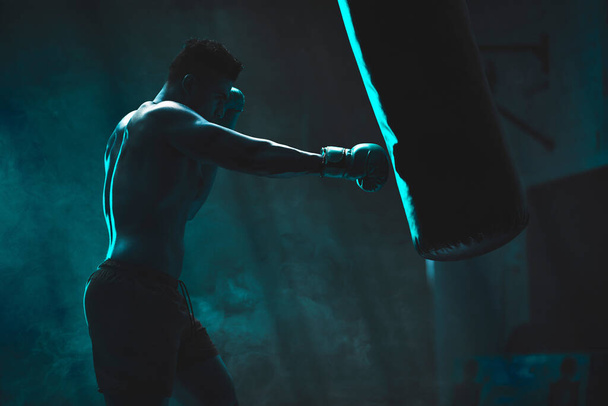 Σιλουέτα ενός αγνώριστου πυγμάχου που παλεύει με σάκο του μποξ σε σκοτεινό γυμναστήριο. Ο αθλητής γυμνάζεται για να προετοιμαστεί για τον επερχόμενο αγώνα του. Εκπαίδευση και εξάσκηση νέων πολεμικών καλλιτεχνών. - Φωτογραφία, εικόνα