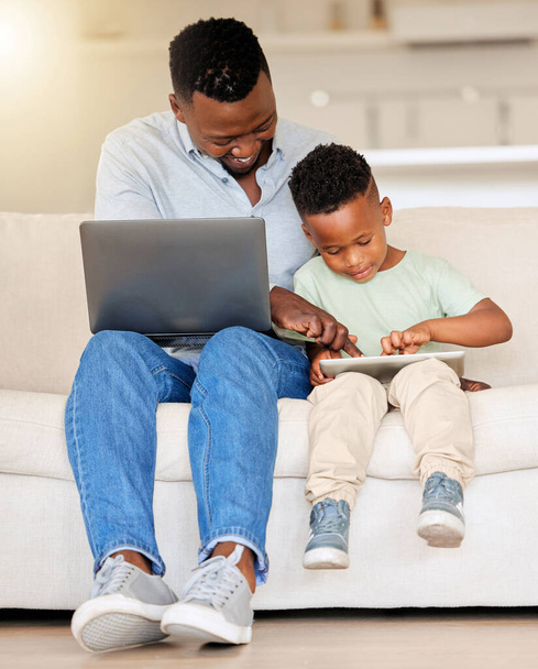 Onnellinen iloinen nuori afrikkalainen amerikkalainen mies käyttää kannettavaa tietokonetta ja auttaa poikaansa digitaalisen tabletin kanssa istuu sohvalla kotona. Isä työskentelee ja lähettää sähköpostia ollessaan yhteydessä poikaansa. - Valokuva, kuva