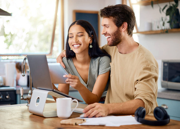 Happy young interracial couple calculating their budget using wireless devices at home. Smiling young caucasian man working on laptop while hispanic wife browses social media on digital tablet. - 写真・画像