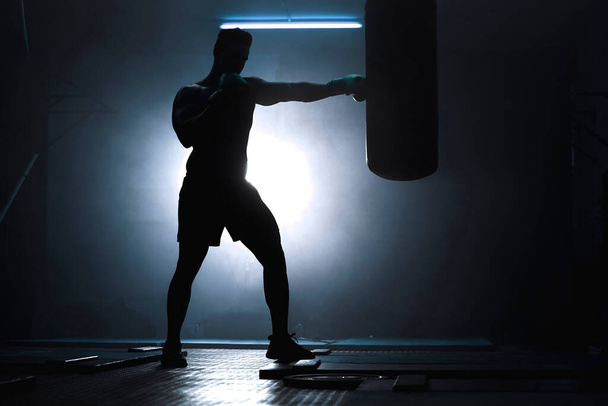 Silhouet van een onherkenbare bokser die spartelt met een bokszak in een donkere sportschool. Mannelijke atleet aan het trainen ter voorbereiding op zijn komende gevecht. Opleiding en training van jonge krijgskunstenaars. - Foto, afbeelding