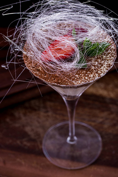 Toetje tiramisu in een glas met aardbeienschijfjes, munt en suikerdraden op een houten ondergrond. Verticale oriëntatie - Foto, afbeelding