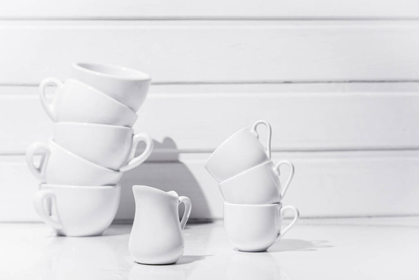 Cups for coffee and tea standing on top of each other and jug for cream on white table and white wood background - Fotoğraf, Görsel