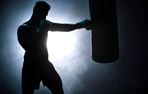 Silhouette of an unrecognizable boxer man sparring with a punching bag in a dark gym. Male athlete working out in preparation for his upcoming fight. Young martial artist training and practicing. - Photo, image