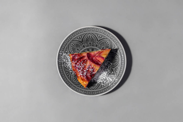 A triangular piece of plum cake with plum slices on top. The cake is sprinkled with sugar pudding. A piece of cake lies on a gray, round, ceramic plate with a pattern. The plate stands on a gray paper background. - Photo, Image