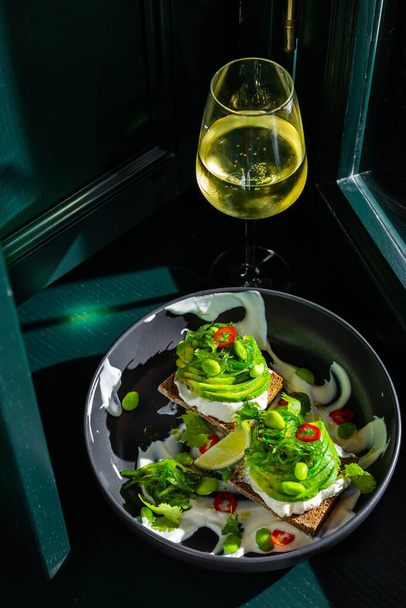 Sandwiches with avocado, beans, chukoy, philadelphia cheese, parsley, pepper and white sauce in a black plate against the background of a dark green window with a glass of wine - Foto, afbeelding