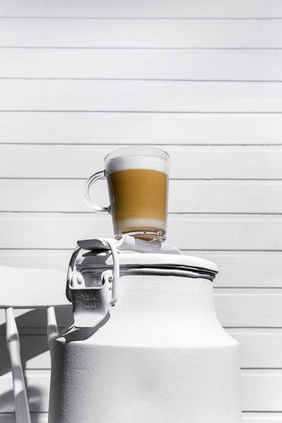 Glass of cappuccino with froth on a white can on a white chair against a background of a white wooden wall - Fotó, kép