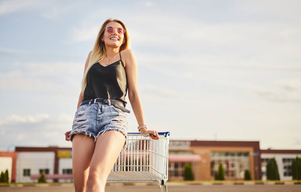 Low angle of glad blond woman in shorts and top with sunglasses smiling and looking away, while leaning on shopping trolley on blurred background of cloudy sky and mall - Photo, image