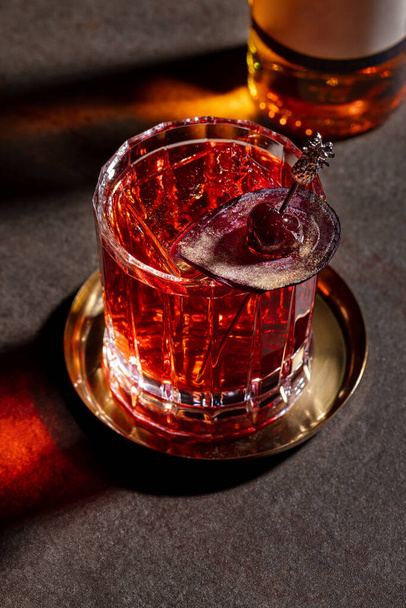 Un vaso de whisky con hielo, cerezas en un pincho y remolachas que se coloca sobre una mesa oscura con una botella de bebida vertida - Foto, imagen
