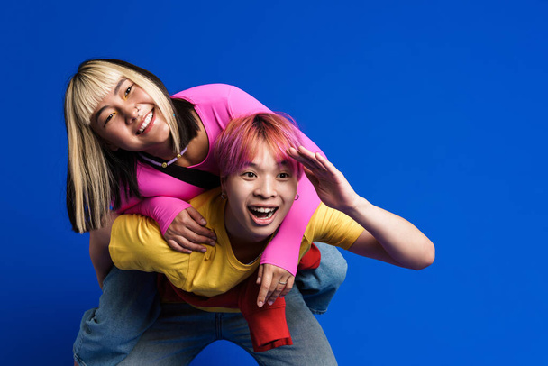 Asian teenagers with multicolored hair laughing while piggybacking isolated over blue wall - Photo, image