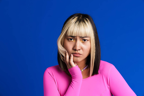 Asian girl with piercing frowning and looking at camera isolated over blue wall - 写真・画像