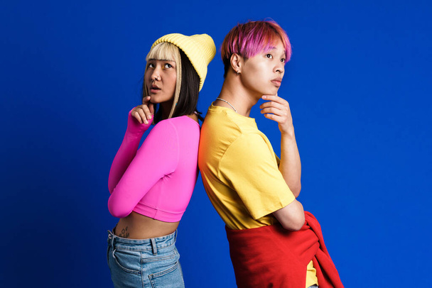 Asian puzzled couple with multicolored hair posing back to back isolated over blue wall - Foto, Imagen