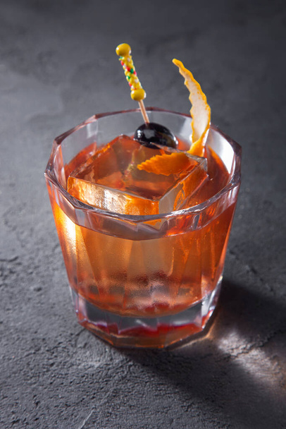 Un cocktail con ghiaccio, un'oliva su uno spiedo e una buccia d'arancia in un bicchiere che poggia su un tavolo di cemento - Foto, immagini
