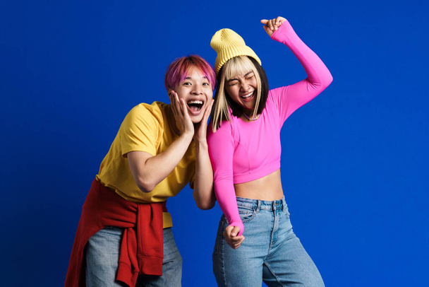 Asian couple with multicolored hair laughing and dancing together isolated over blue wall - Fotó, kép