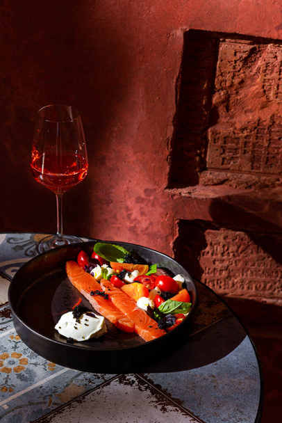 Салат з філе лосося, помідорами, базиліком, сиром фета, ікрою та соусом у тарілці на столі з келихом вина на коричневій стіні
 - Фото, зображення
