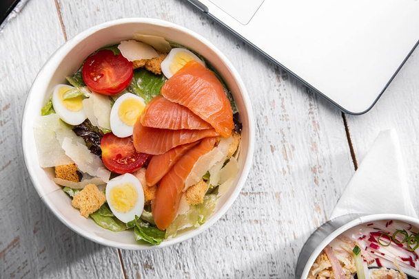 Salad with salmon, egg, tomatoes, croutons, cheese and salad in a paper box standing on a table with a laptop - Photo, image
