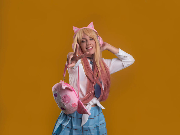 Content female model in kitty headphones with pink handbag showing two fingers gesture and looking at camera against brown background - Foto, Bild