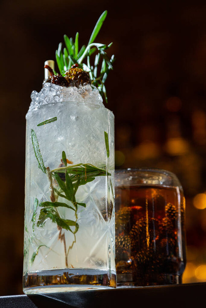 A cocktail with ice, rosemary, small cones and a straw in a glass that stands on the table against the background of burning lights - Valokuva, kuva