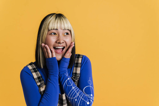Asian girl with piercing laughing and looking aside isolated over yellow wall - Фото, изображение
