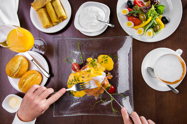 Breakfast at the hotel from egg benedict with salmon, cheese sauce, cherry tomatoes, microgreen in a glass, transparent plate. Male hands are holding a fork with a knife. To the right of the plate is a cup of cappuccino. Above is a white plate of qua - Photo, image