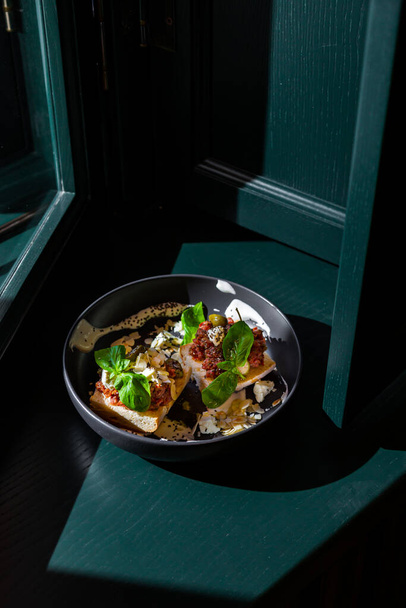 Sandwiches with cheese, meat, basil, almonds, kumquat, sauce and sesame seeds in a black plate against a dark green window background - Foto, Bild