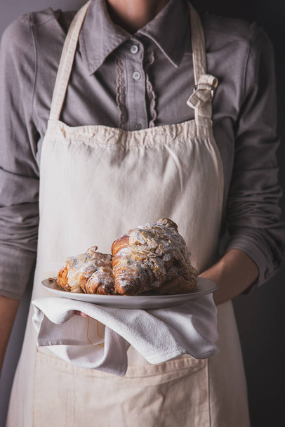 A girl in an apron and a shirt holding croissants with condensed milk, powdered sugar and slices of almonds with a towel on a plate - Zdjęcie, obraz