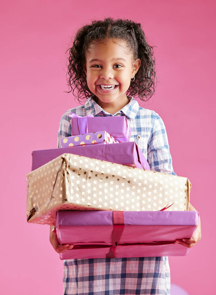 Ładna mieszanka rasy dziewczyna z kręconymi włosami trzymająca stos zapakowanych prezentów na różowym tle copyspace w studio. Afrykańskie dziecko wyglądające podekscytowane dostaniem prezentów na urodziny. - Zdjęcie, obraz