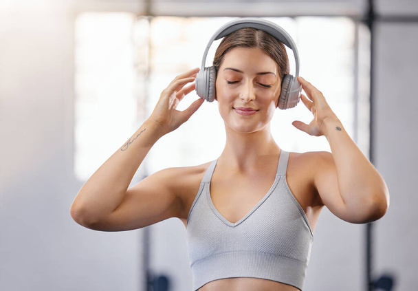One active young caucasian woman listening to relaxing music with headphones while taking a break from exercise a gym. Female athlete staying motivated with calm music during her workout in a fitness - Photo, Image