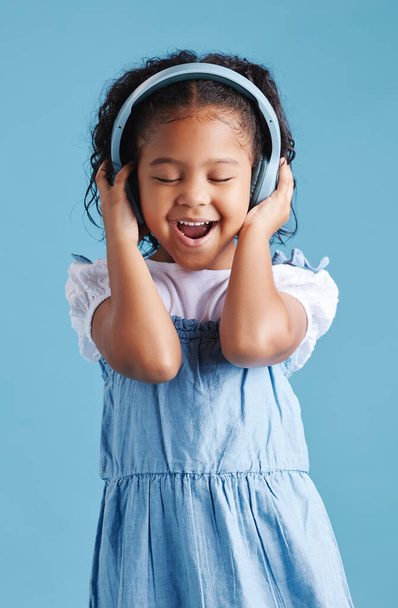 Adorable little hispanic girl standing with her eyes closed and looking happy while wearing wireless headphones and enjoying her favourite song against a blue studio background. - Photo, image