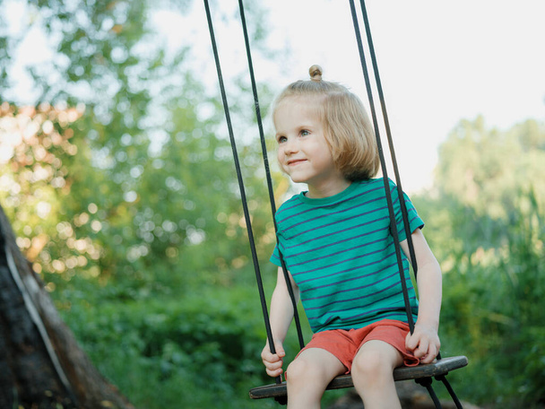 Adorable little boy with long blond hair swings on a fresh air on a sunny summer day. Child wearing green t-shirt and red shorts. - Photo, image