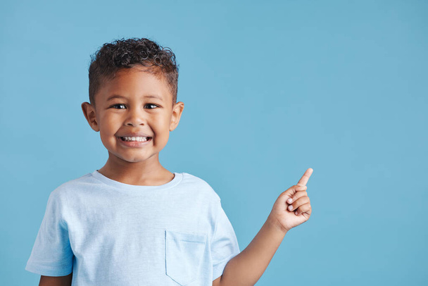 Portret van een vrolijk lachend jongetje dat met zijn vinger wijst naar kopieerruimte naar rechts tegen een blauwe studioachtergrond. Vrolijk Latijns-Amerikaans kind met gezonde tandheelkundige glimlach in casual kleding. - Foto, afbeelding