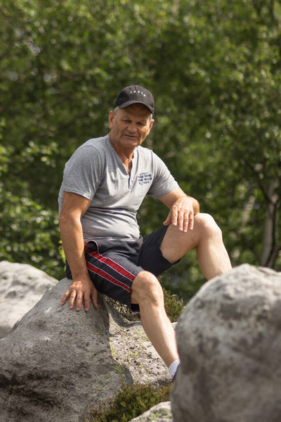 Lächelnder kaukasischer Senior im grauen T-Shirt mit schwarzer Baseballkappe. Nachdenklicher alter Mann sitzt auf dem Stein und ruht sich aus, nachdem er oben auf dem Berg gewandert ist. Natur im Hintergrund. Aktive Senioren. - Foto, Bild
