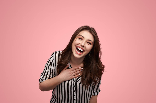 Joyful young female millennial with long brown hair in striped shirt laughing happily and looking at camera against pink background - Photo, Image