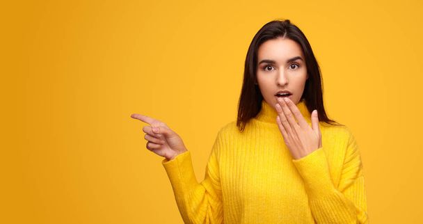 Astonished young Hispanic female in sweater covering mouth and looking at camera while pointing at empty space during advertisement campaign against yellow background. - Foto, Imagen