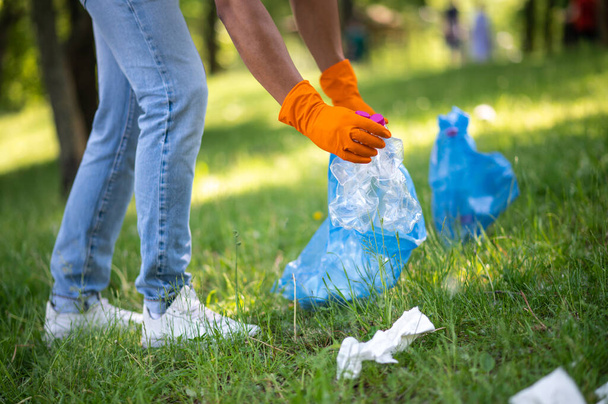 Care. Gloved hands of dark-skinned man putting plastic waste into bag on grass in park on sunny day without face - Фото, изображение