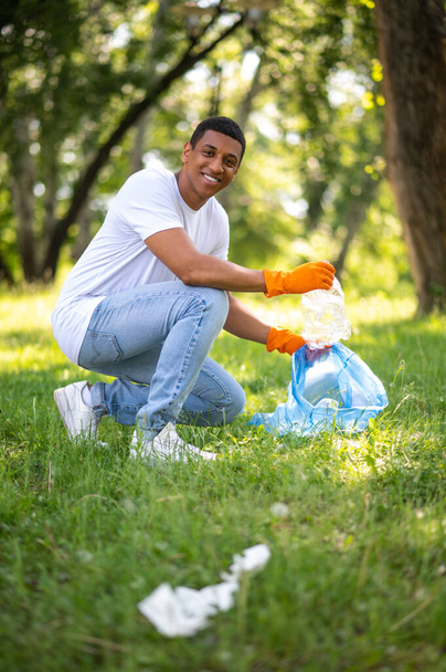 No plastic. Young dark-skinned man crouched sideways to camera picking up plastic waste smiling at camera in park - 写真・画像
