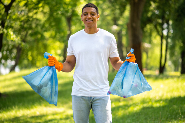 Good result. Young dark-skinned man in gloves smiling at camera holding garbage bags standing in nature on sunny day - Fotografie, Obrázek