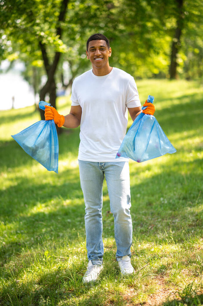 Volunteering. Young dark-skinned guy with bag standing holding garbage bags in park smiling confidently at camera - Fotografie, Obrázek