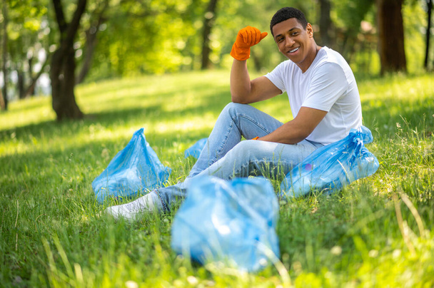 Щасливий день. Усміхнений темношкірий хлопець в захисних рукавичках сидить біля пакетів, що відпочивають на траві в парку
 - Фото, зображення