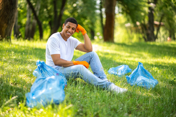 Resultaat van het werk. Donkerhuid man in beschermende handschoenen zittend op gras glimlachend kijkend naar vuilnis verzameld in zakken in park - Foto, afbeelding