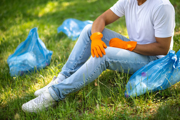 Volunteering. Dark-skinned man in protective gloves resting sitting on grass near garbage bags on sunny day no face - Foto, Imagen